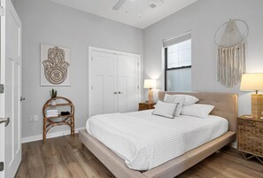 A bedroom with a large bed, a white door, a wall-mounted macrame decoration, and a window with blinds.