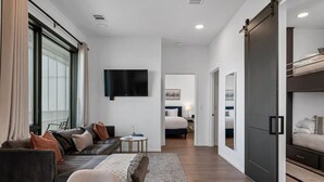 A modern living room with a sofa, coffee table, and a TV. There is a bedroom with a bed and a bunk bed. - GULCH Condo with Pool, Gym, Private Patio & Garage (Nashville)