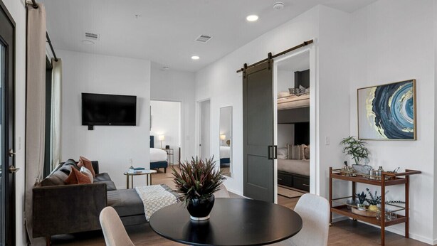 A modern living room with a sofa, a dining table, and chairs. There is a TV mounted on the wall, a sliding door, and a plant on the table. - GULCH Condo with Pool, Gym, Private Patio & Garage (Nashville)