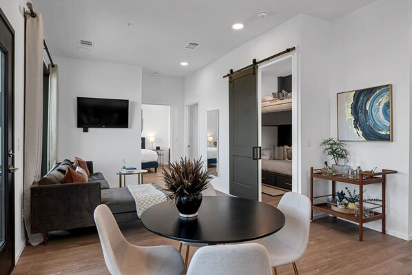 A modern living room with a sofa, a dining table, and chairs. There is a TV mounted on the wall, a sliding door, and a plant on the table.