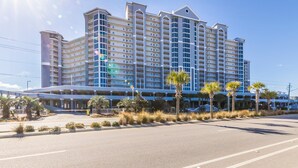 A multi-story hotel with a central atrium, palm trees, and a parking lot. - Lighthouse 402 | Beachfront | Waterspouts + Zero Entry Pool | My Beach Getaways (Gulf Shores)
