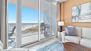 A room with a balcony, a sofa, a coffee table, and a painting on the wall. - Lighthouse 402 | Beachfront | Waterspouts + Zero Entry Pool | My Beach Getaways (Gulf Shores)
