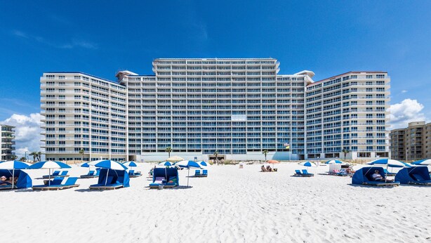 A large hotel with multiple floors and balconies, a sandy beach in the foreground, and several beach umbrellas and lounge chairs. - Lighthouse 402 | Beachfront | Waterspouts + Zero Entry Pool | My Beach Getaways (Gulf Shores)