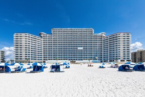 A large hotel with multiple floors and balconies, a sandy beach in the foreground, and several beach umbrellas and lounge chairs. - Lighthouse 402 | Beachfront | Waterspouts + Zero Entry Pool | My Beach Getaways (Gulf Shores)