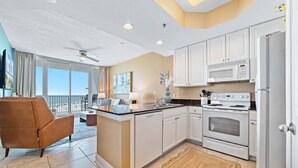 A modern kitchen with white cabinets, a built-in microwave, and an oven. There is a brown armchair and a dining area with a view of the outdoors. - Lighthouse 402 | Beachfront | Waterspouts + Zero Entry Pool | My Beach Getaways (Gulf Shores)