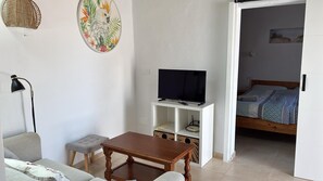 A room with a wooden table, a sofa, a television on a white stand, and a bed with a patterned bedspread.