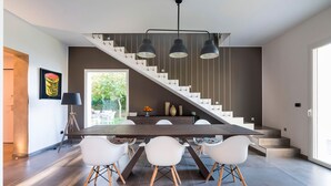 A modern kitchen with a large dining table, white chairs, and a staircase with pendant lights.