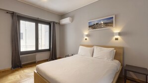 A bedroom with a large bed, a window with curtains, an air conditioning unit, a bedside table, and a wall-mounted picture. - Heyden 3 · Top Floor Renv 3BDR APT / “Pedion tou (Athens)