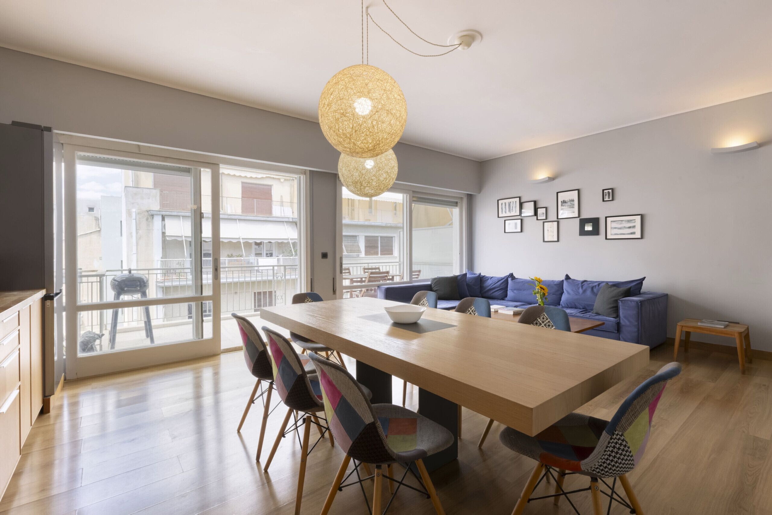 A modern living room with a wooden dining table, colorful chairs, a blue sofa, and a large window offering a view of buildings outside.