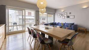A modern living room with a wooden dining table, colorful chairs, a blue sofa, and a large window offering a view of buildings outside. - Heyden 3 · Top Floor Renv 3BDR APT / “Pedion tou (Athens)