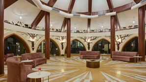 A spacious lobby with high ceilings, large arched windows, and a central seating area with sofas and round tables. - The Sawai Bagh (Sawai Madhopur)