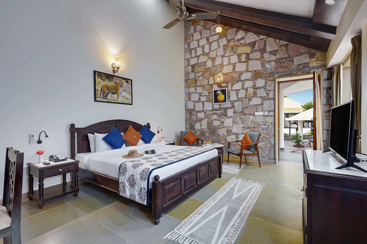 A bedroom with a stone wall, a bed with patterned bedding, a nightstand, a chair, a TV, and a view of the outside.