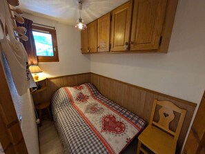 Um quarto pequeno e acolhedor com armários de madeira, cama com manta estampada, uma mesa pequena e uma janela com cortinas.