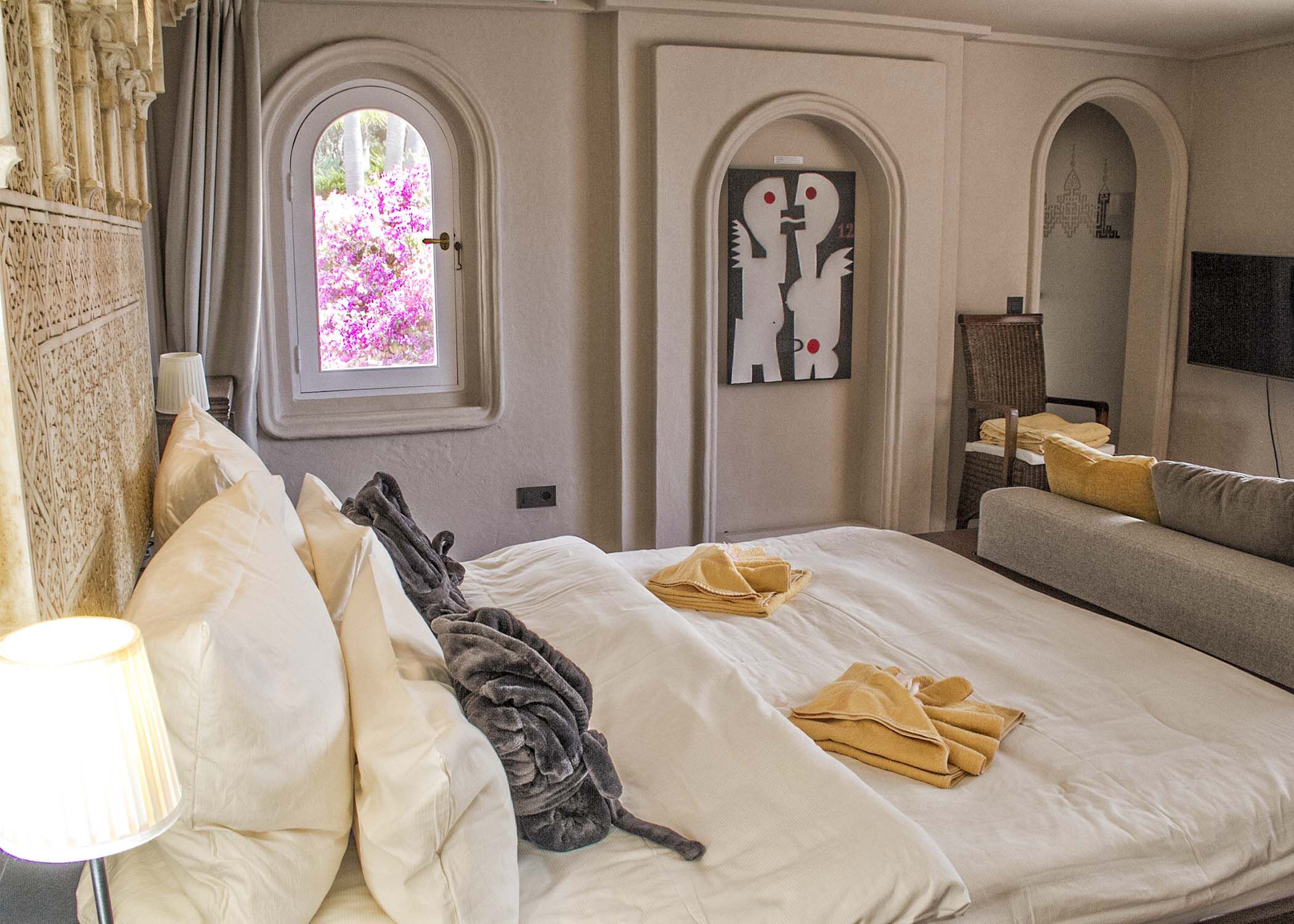 A hotel room with a large bed, a sofa, a TV, and a window with a view of pink flowers.