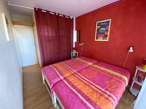 A bedroom with a bed, a red wall, a painting, and a small table with a lamp.