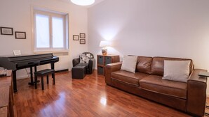 A living room with a piano, a brown leather sofa, a small table with a lamp, and a window with blinds. - Comfortable tastefully furnished apartment in a strategic position (Bergamo)