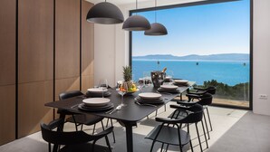 Comedor moderno con una gran mesa dispuesta para seis comensales, vista al mar y un panel de madera en la pared.