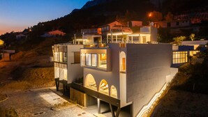 Un moderno edificio de varios pisos con amplios ventanales y balcones, ubicado en una colina y rodeado de viviendas.