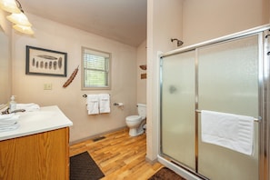 Een badkamer met een houten wastafelmeubel, een glazen douchecabine, een toilet en een lijst met een veer erin.
