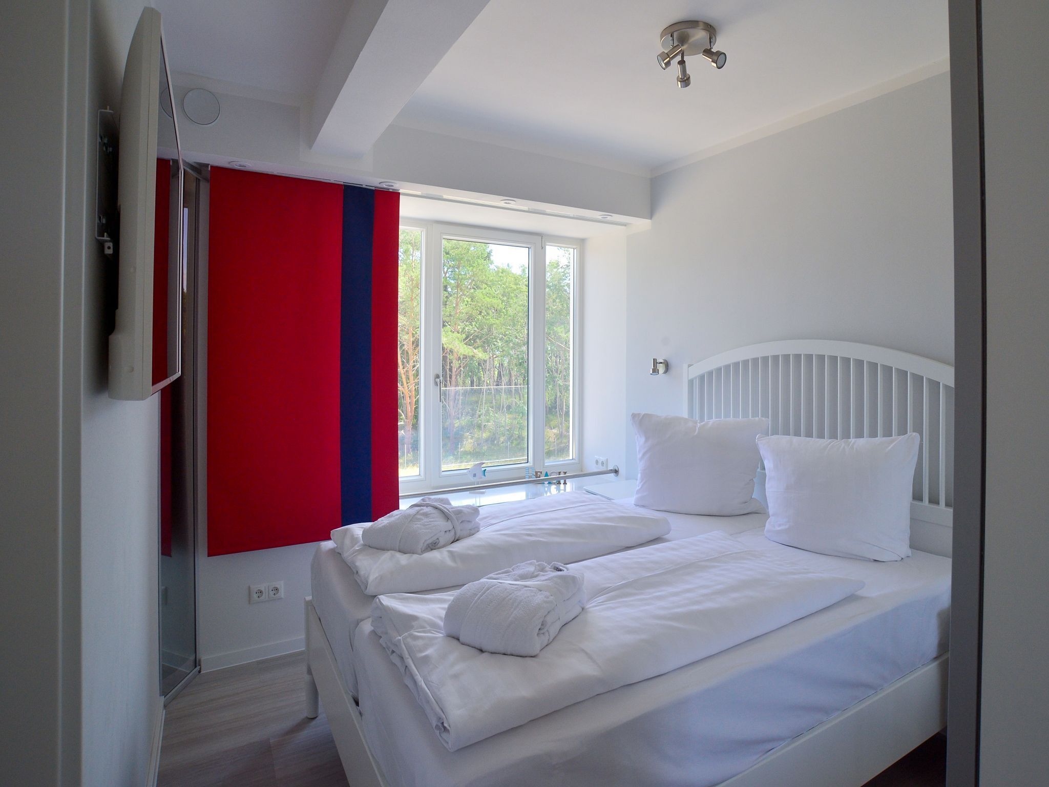 Une chambre avec un lit blanc, une fenêtre donnant sur des arbres et un rideau rouge et bleu.