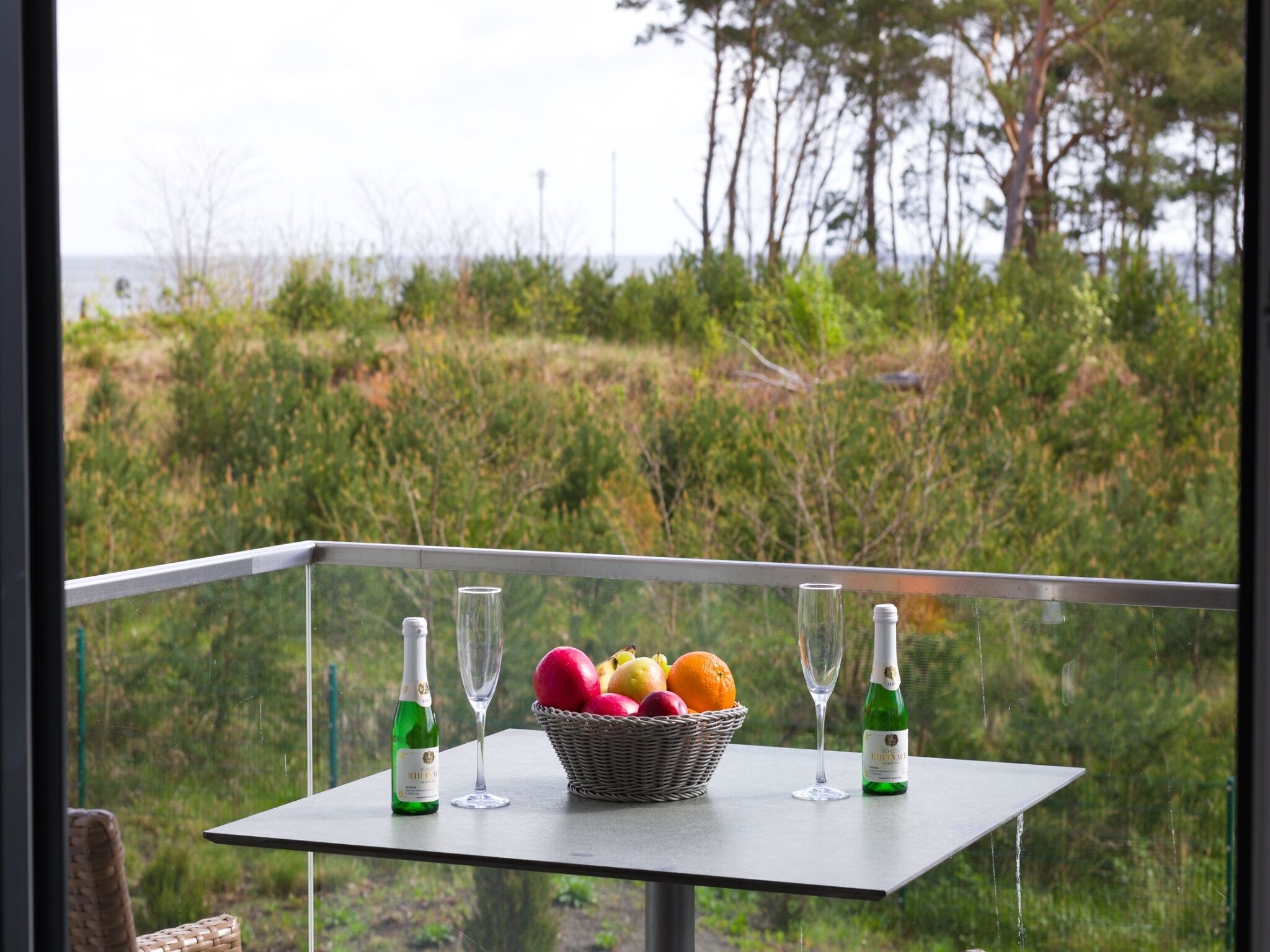 Una mesa con una cesta de frutas, dos botellas y dos vasos en un balcón con vistas a zonas verdes y árboles.