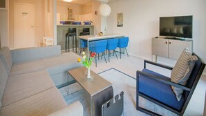 A modern living room with a sofa, a coffee table with a vase, a TV, and a dining area with blue chairs. - Apartment with sea view, 2 bedrooms and balcony (Ostseebad Binz)