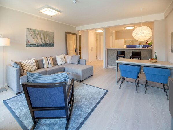 A modern living room with a grey sectional sofa, a dining area with blue chairs, and a kitchen in the background.