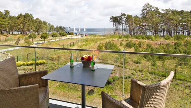 Un balcon agrémenté de fauteuils et d’une table en osier, offrant une vue sur un paysage côtier avec des arbres et la mer.