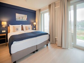 A hotel room with a large bed, two bedside tables with lamps, a painting on the wall, and a view through the window. - Apartment with a view of the ocean, 3 bedrooms, and a balcony (Ostseebad Binz)
