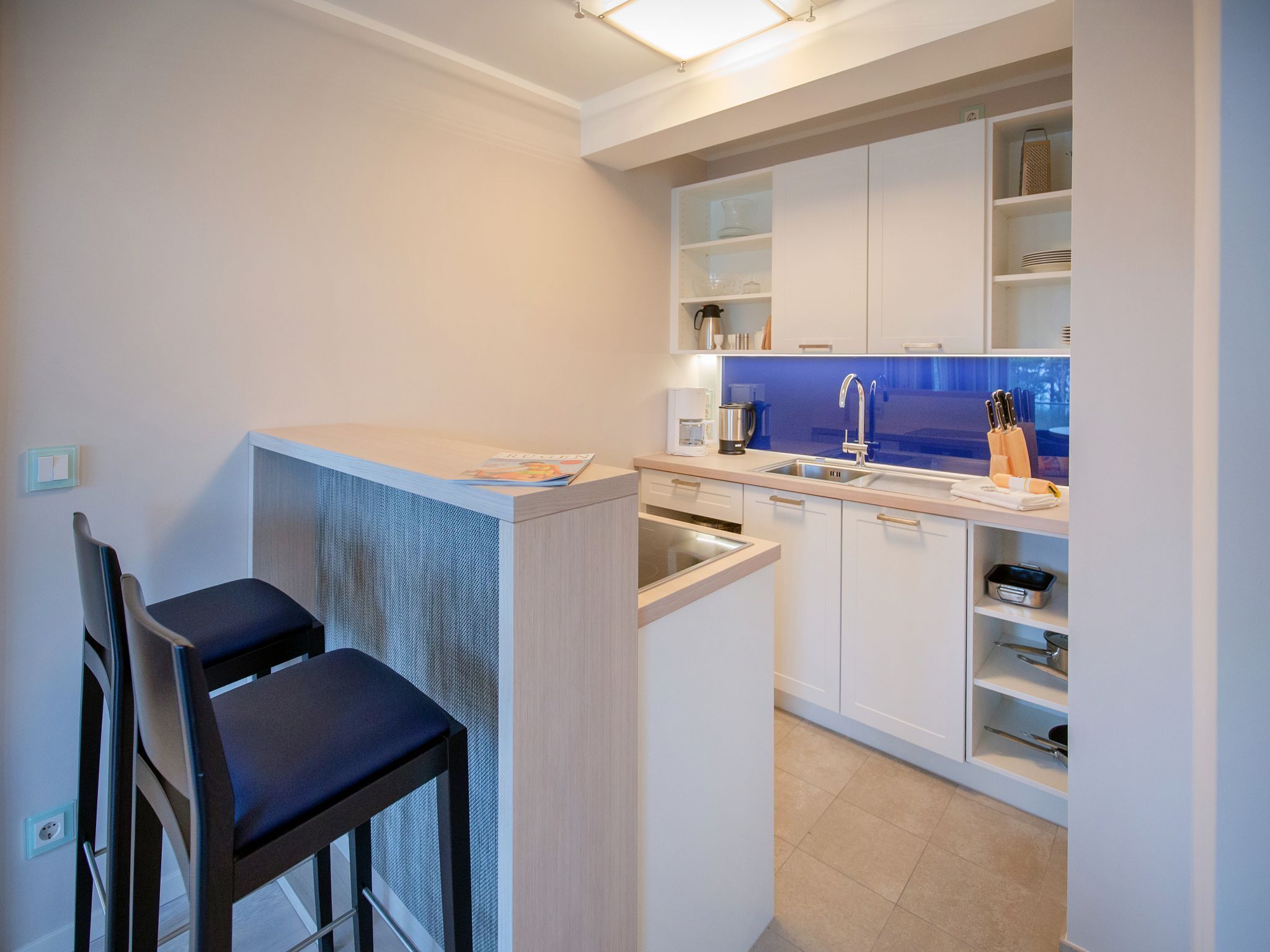 A compact kitchen with white cabinets, a sink, and a coffee machine. There are two chairs and a small table.