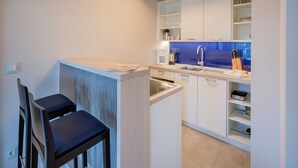 Cocina compacta con gabinetes blancos, un lavamanos y una cafetera. Hay dos sillas y una pequeña mesa.
