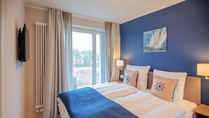 Habitación de hotel con una cama grande, un cuadro de un velero y vistas a los árboles desde la ventana.