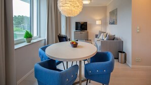 Una sala de estar moderna con una mesa de comedor redonda, sillas azules, una lámpara colgante blanca, un televisor y un sofá gris.