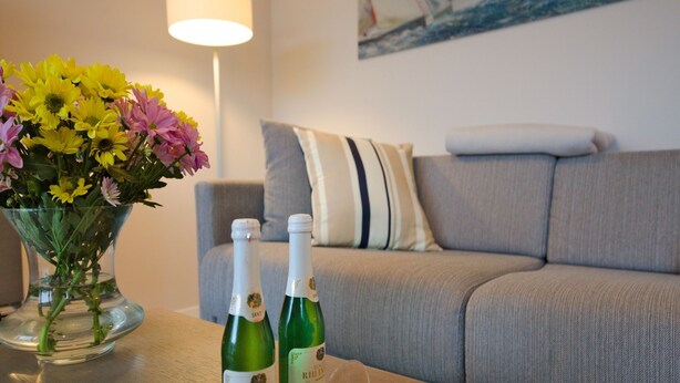 Una sala de estar con un sofá, un florero con flores, dos botellas de vino y una copa de vino sobre una mesa de madera.