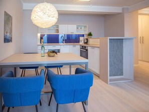A modern kitchen with white cabinets, a dining table with blue chairs, and a hanging light fixture. - Apartment with a view of the sea, 1 bedroom, and a balcony (Ostseebad Binz)