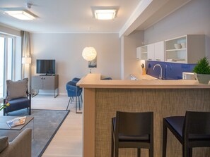 A modern kitchen with a central island, a dining area with chairs, a sofa, and a TV in the background. - Apartment with sea view, 1 bedroom and balcony (Ostseebad Binz)