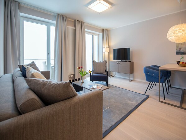 A modern living room with a sofa, a dining table, a TV, and a view of the outdoors. - Apartment with a view of the sea, 1 bedroom, and a balcony (Ostseebad Binz)