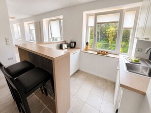 A modern kitchen with a central island, a dining area with a black chair, and a window with a view of greenery. - Apartment with a view of the sea, 1 bedroom, and a balcony (Ostseebad Binz)