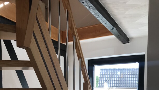A wooden staircase with a black railing, a window with white vases, and a wooden cabinet with a bottle. - Duplex apartment in a conservation area (Solingen)