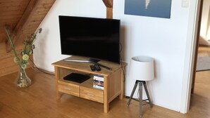 A cozy room with a wooden ceiling, a flat-screen TV on a wooden TV stand, a small white lamp, and a painting of a sailboat on water. - Duplex apartment in a conservation area (Solingen)
