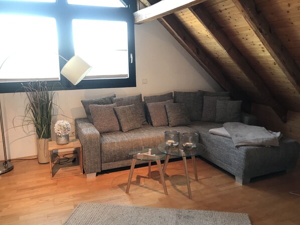 A cozy loft-style room with a wooden ceiling, a grey sofa with multiple cushions, a small glass table with two glasses, and a potted plant. - Duplex apartment in a conservation area (Solingen)