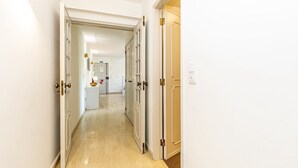 En gang med hvide vægge og døre, der fører til et køkkenområde med et køleskab og en vask.