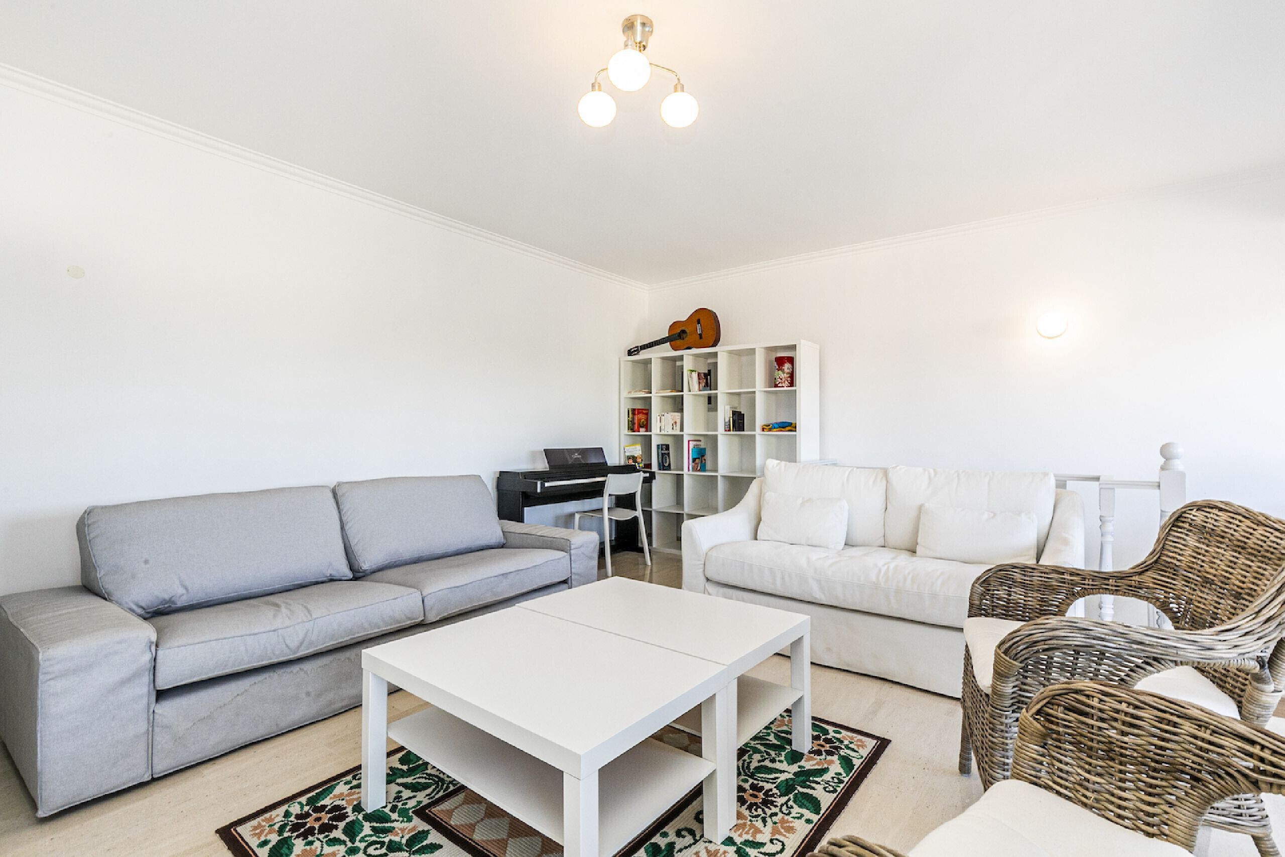 Ein Wohnzimmer mit grauem Sofa, weißem Sofa, weißem Couchtisch, Korbstühlen, einem Bücherregal und einer Deckenleuchte.