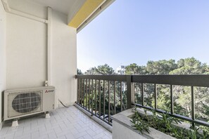 A balcony with a Mitsubishi air conditioning unit, a concrete planter with plants, and a view of trees and buildings in the background.