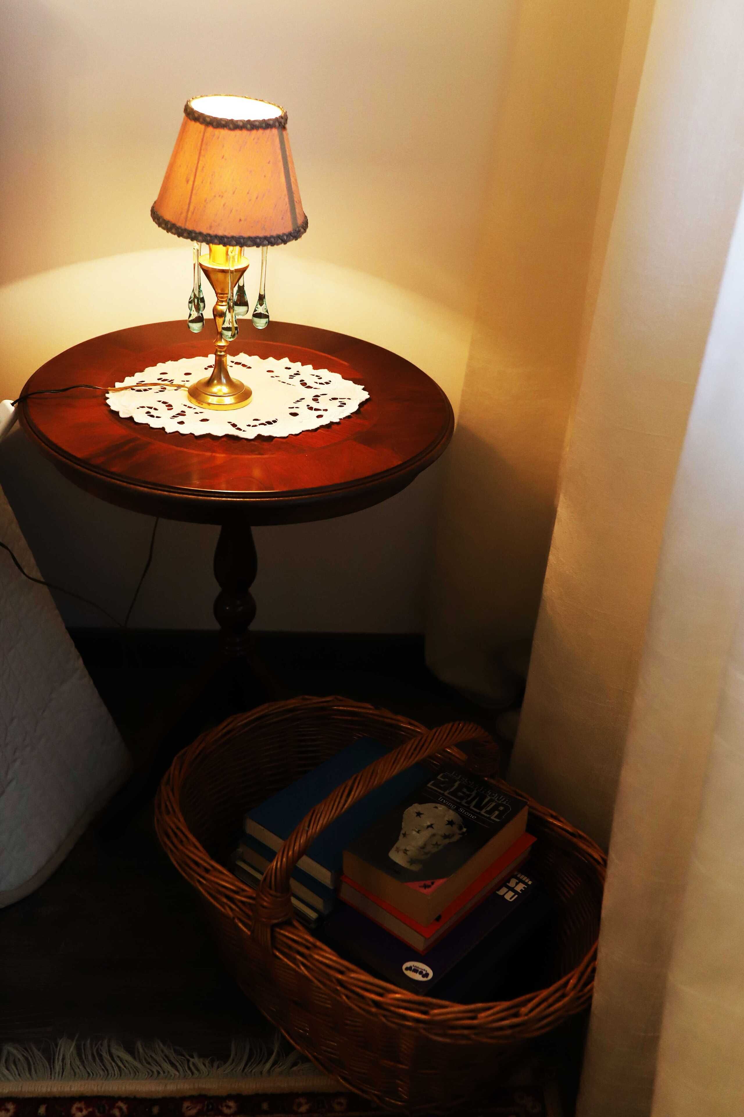 A corner with a wooden table, a lit lamp, and a wicker basket containing books.