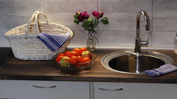 Una cocina con un lavamanos de acero inoxidable, una cesta de mimbre, un cuenco de fruta y un jarrón con flores.