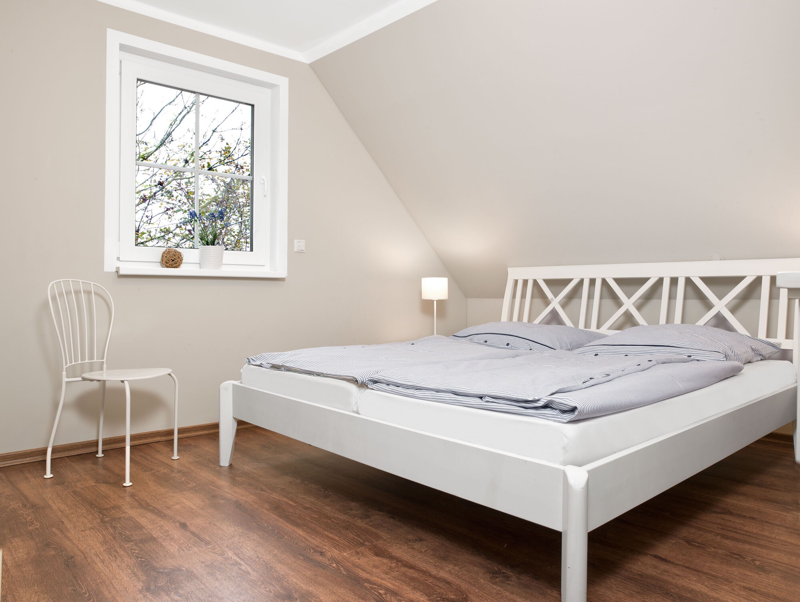 Une chambre avec un lit blanc, une petite chaise blanche, une fenêtre donnant sur des arbres et une lampe sur la table de chevet.