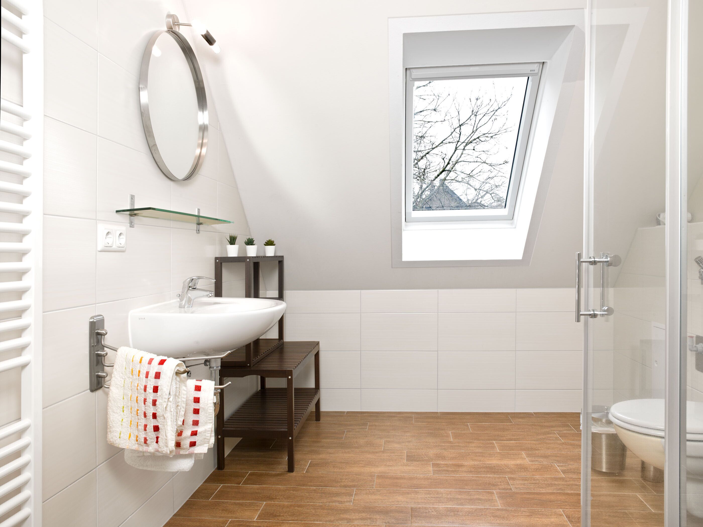 Un baño moderno con tragaluz, un lavamanos blanco, cabina de ducha de vidrio y suelo de madera.
