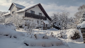 A house with a sloped roof and a wooden balcony, surrounded by snow-covered trees and a snow-covered ground.