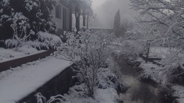 A snow-covered landscape with a building featuring a covered walkway and a small stream.